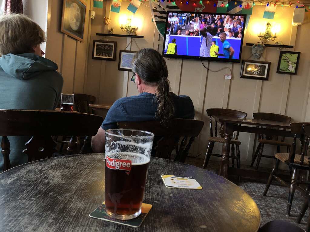 Ein Pint Bier auf einem Tisch in einem Pub, im Hintergrund ein TV in dem Fußball läuft