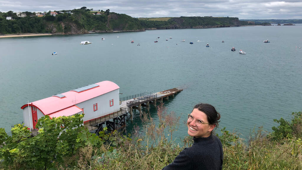 Eli steht auf einer Klippe überm Meer, darunter ein Bootshaus mit schräg ins Meer laufendem Steg