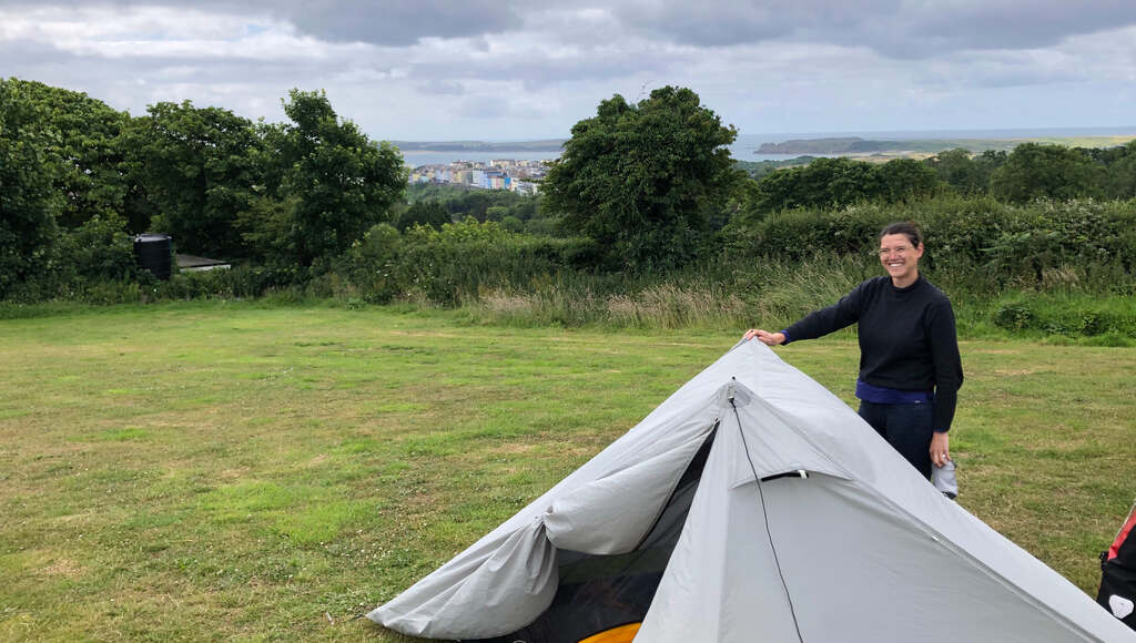 Kathi steht am Zelt auf einer Wiese, am Horizont Meer, Klippen und ein Ort