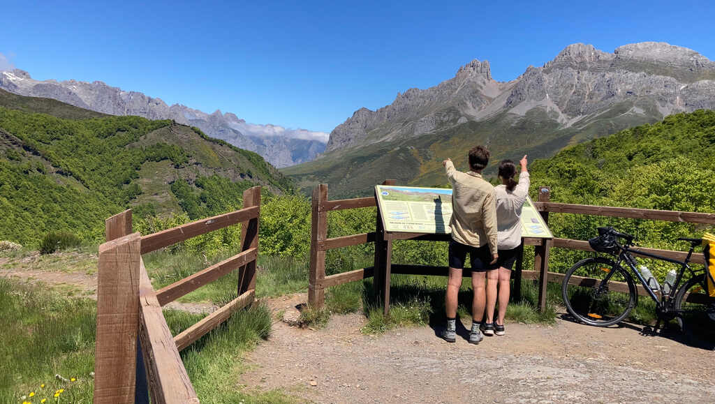 Kathi und Eli stehen an einer Infotafel vor einem Bergpanorama und zeigen in unterschiedliche Richtungen