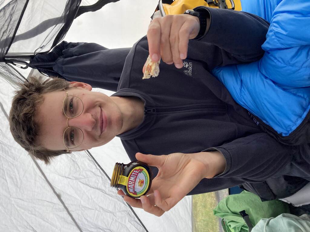 Eli sitzt im Zelt und zeigt ein Stück Brot und einen Tiegel Marmite