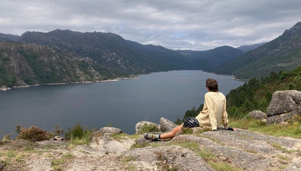 Eli sitzt auf einem Aussichtspunkt über einem See zwischen Bergen