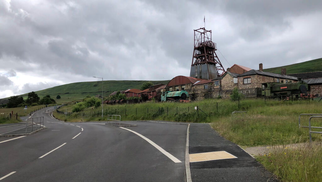 Ein Straße führt vorbei an einem Gelände mit alten rot lackierten Industriegebäuden und einem hohen Turm mit einer großen Spule