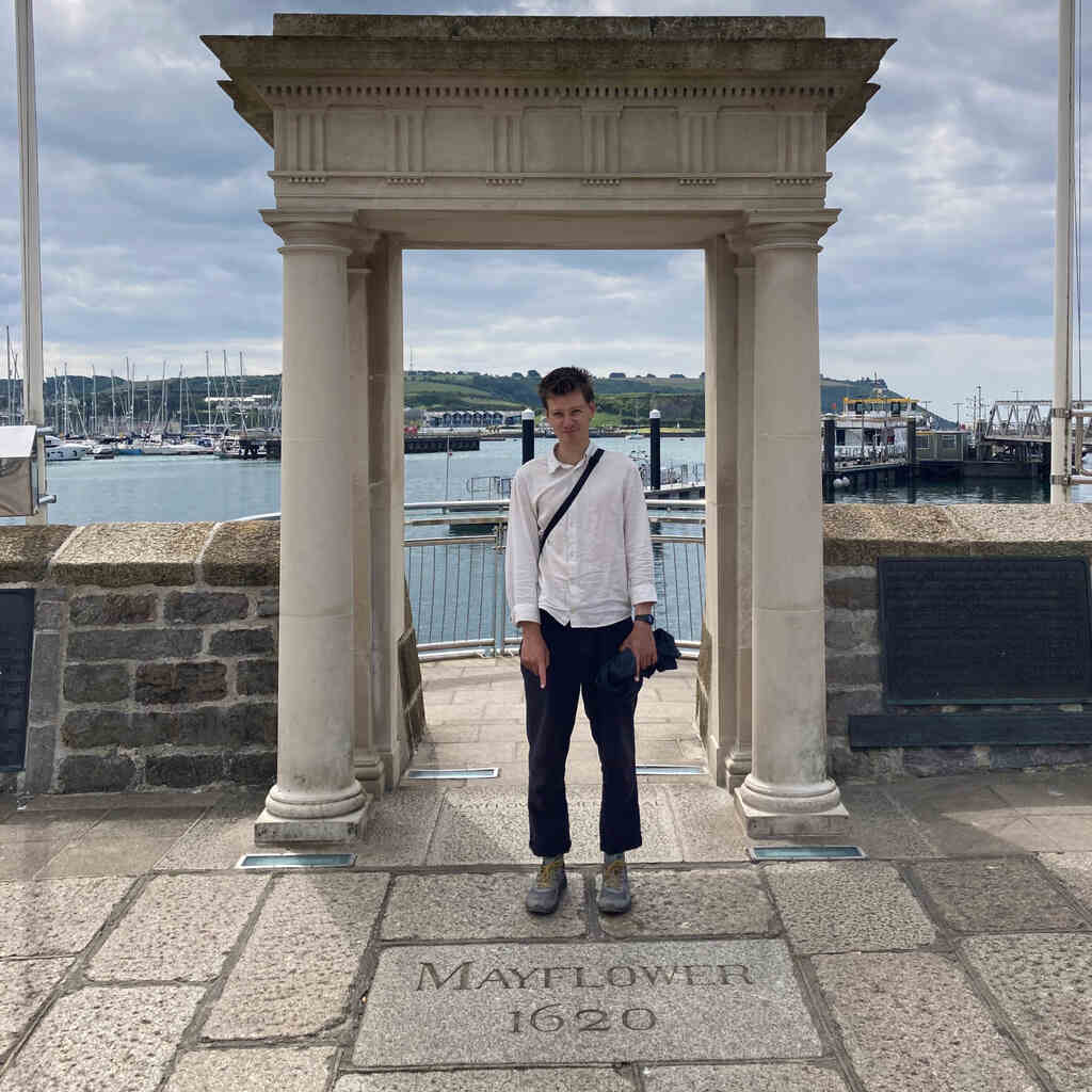 Eli steht unter einem Portico und zeigt auf eine Gedenktafel zur Mayflower im Boden