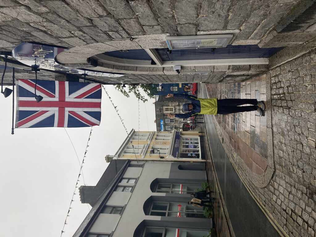 Eli steht auf der Straße mit Regenjacke und Kapuze und zeigt auf den Union Jack der über ihm an der Fassade hängt