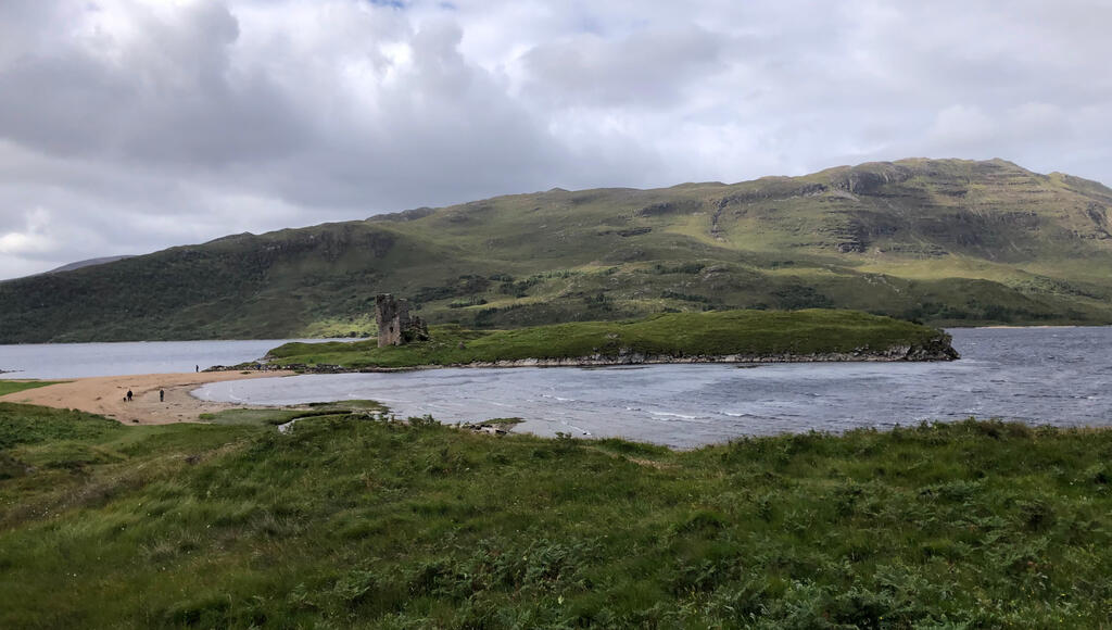 Eine grasige Halbinsel verbunden mit dem Festland über einen Sandstrand, auf der Insel die Ruine einer kleinen Burg