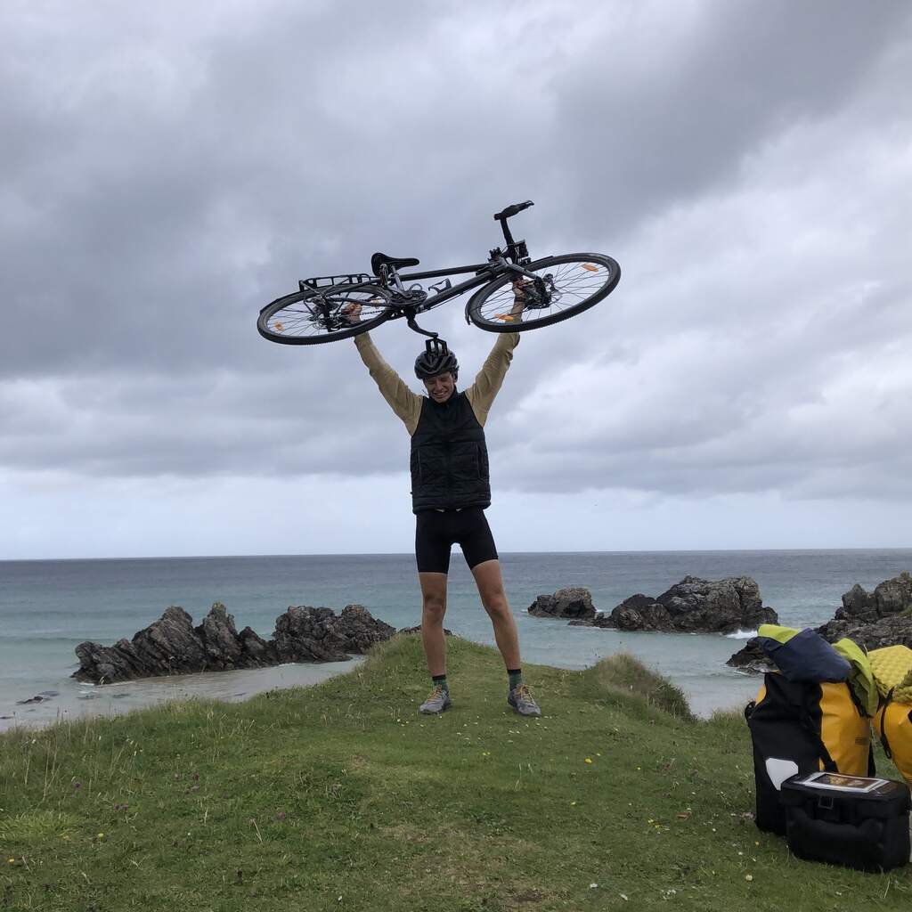 Eli steht auf einer kleinen Wiese über einem felsendurchsetzten Strand und reckt grinsend sein Fahrrad über den Kopf