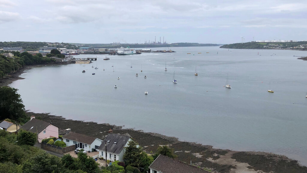 Eine breite Flussmündung mit verstreuten Seegelbooten und einer Raffinerie am Horizont