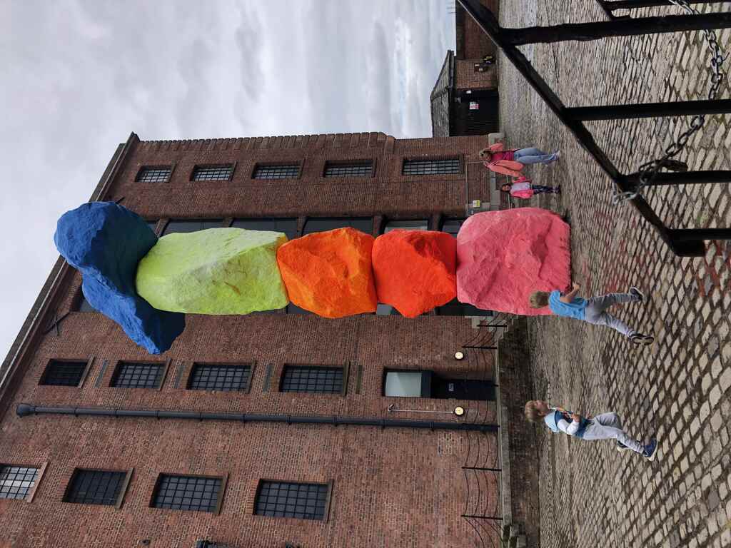 Eine Skulptur aus aufeinandergestapelten bunt bemalten Natursteinen, Kinder laufen drumherum