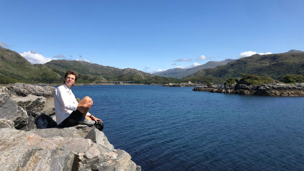 Eli sitzt auf unregelmäßigen Felsblöcken am Wasser, im Hintergrund eine Berglandschaft und kleine felsige Inseln im Meer