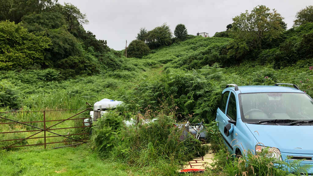 Ein himmelblauer Fiat Panda rechts von einem rostigen Weidetor, dahinter ein steiler Hügel hoch zu einer kleinen Hütte