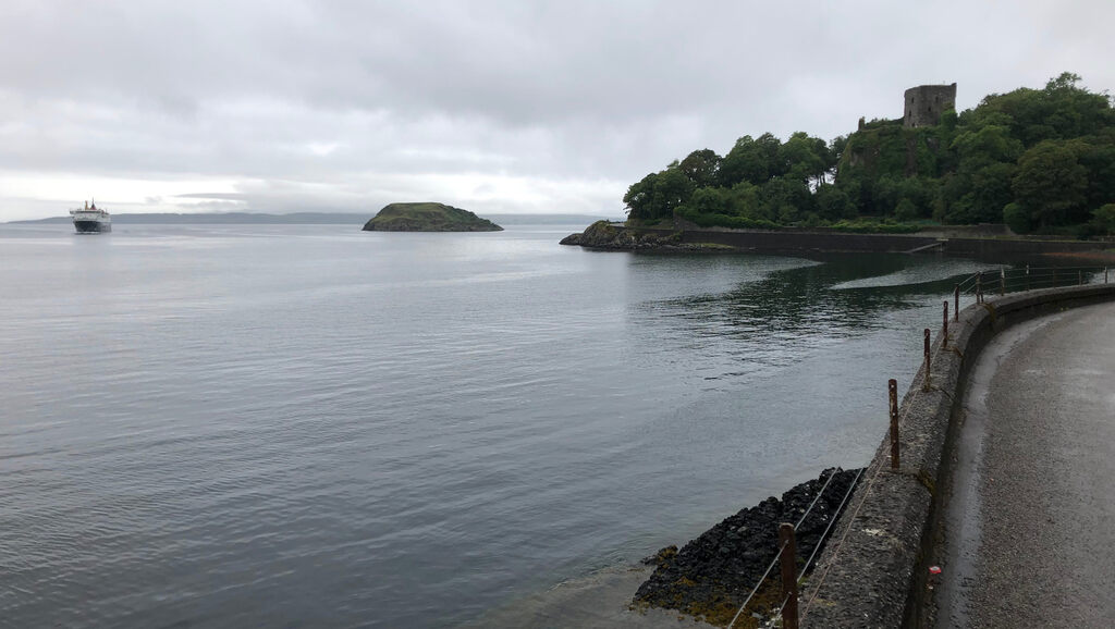 Eine Straße in regnerischer Witterung mit Blick aufs Meer und einer Turmruine auf einem waldigen Hügel, im Meer ein Fährschiff