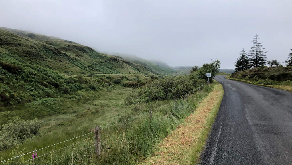 Eine Straße schlängelt sich sanft bergauf, an der linken Seite grasige Hügel deren Spitzen im Nebel liegen