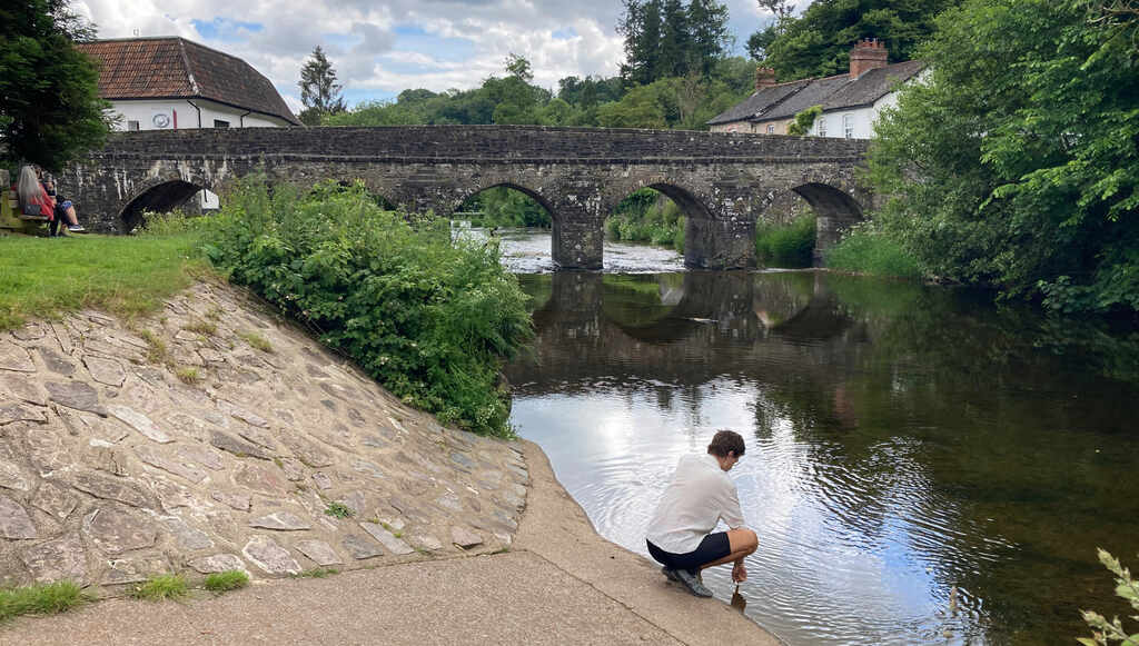 Eli in der Hocke an einem Flussufer, im Hintergrund eine Steinbrücke