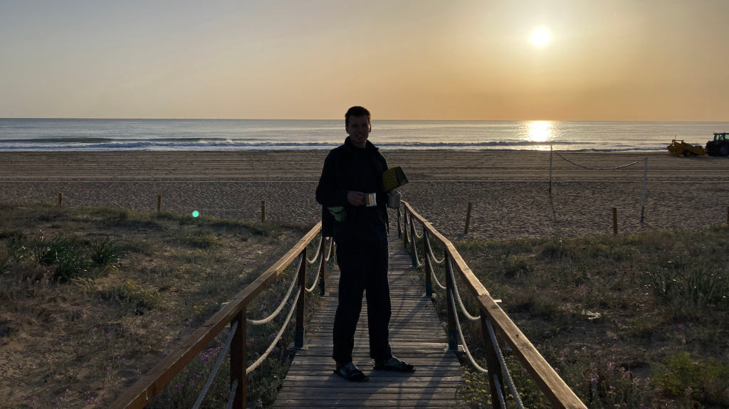 Eli steht mit zwei Kaffeetassen am Strand