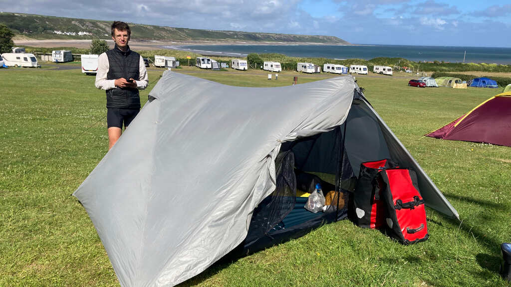 Eli steht vorm aufgebauten Zelt am Campingplatz, dahinter eine Reihe Wohnwägen, noch weiter dahinter eine langgezogene Bucht