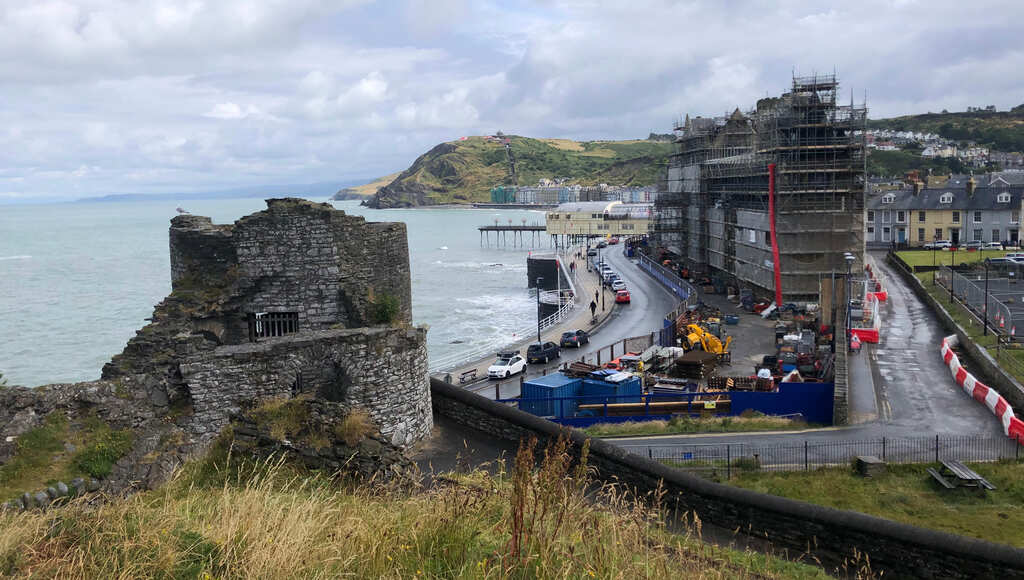 Eine Ruine im Vordergrund, dahinter eine Straße am Meer entlang und ein Gebäude in Gerüste gekleidet, am Horizont ein Hügel