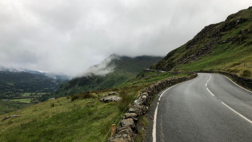 Eine nasse Straße führt auf Kurven den Berg hoch, im Hintergrund wolkenverhangene Hügel