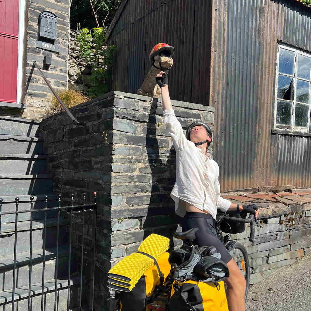 Eli sitzt am Rad und streckt den Arm hoch zu einem holzgeschnitzten Schwein mit einem Kinderfahrradhelm am Kopf