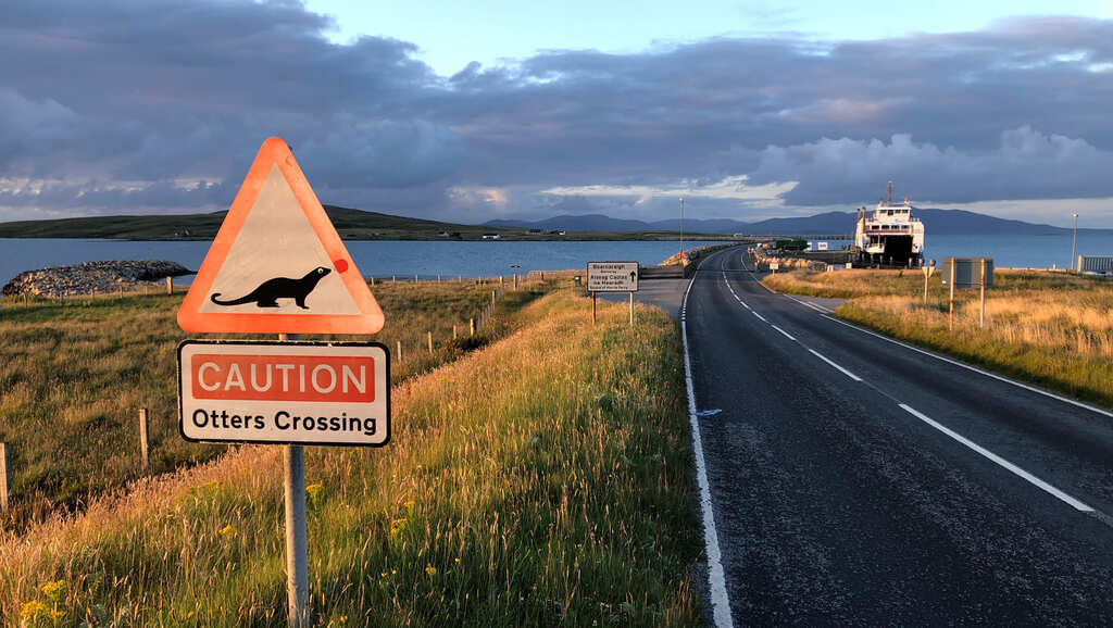 Eine Brücke übers Meer, am Ufer ein Fährschiff, an der Straße ein Schild auf dem steht 'CAUTION Otters Crossing'