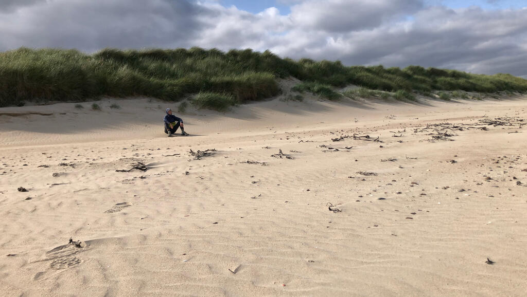 Eli sitzt in Windjacke an einer grasbewachsenen Düne am Strand